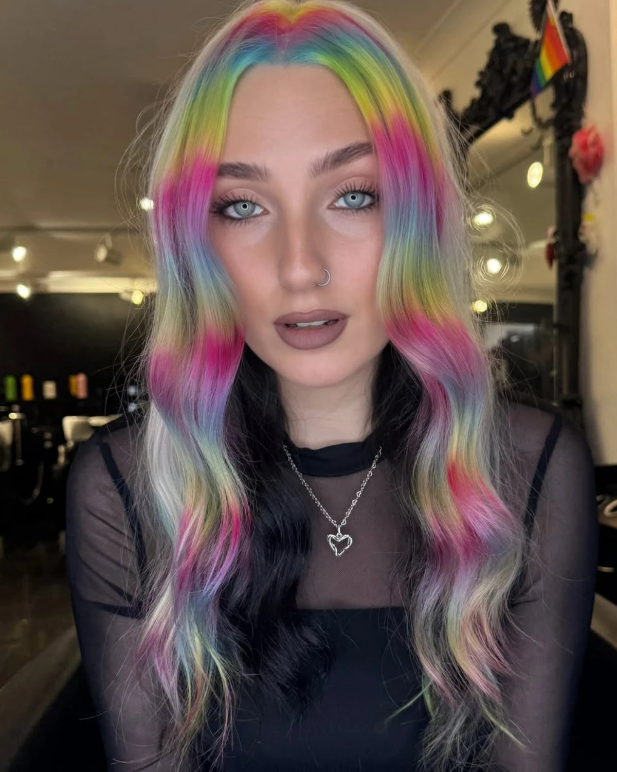A woman with long, wavy rainbow-colored hair in pastel shades of pink, blue, green, yellow, and purple, wearing a black sheer top and a silver heart pendant necklace in what appears to be a salon interior.