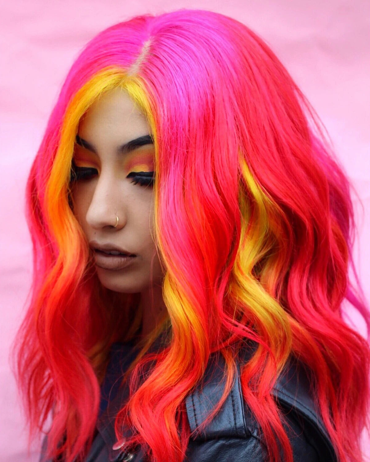 A close-up portrait of a person wearing a vibrant, wavy wig with a bright pink, orange, and yellow gradient. The background is a solid light pink color.