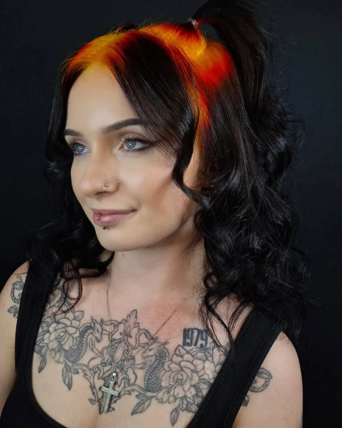 A close-up portrait of a woman with long, wavy black hair featuring a vibrant orange and yellow streak at the crown, wearing a black tank top and displaying floral tattoos on her chest against a dark background.