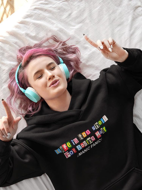 Person wearing a black hoodie with colorful text, lying on a bed with pink hair and teal headphones.